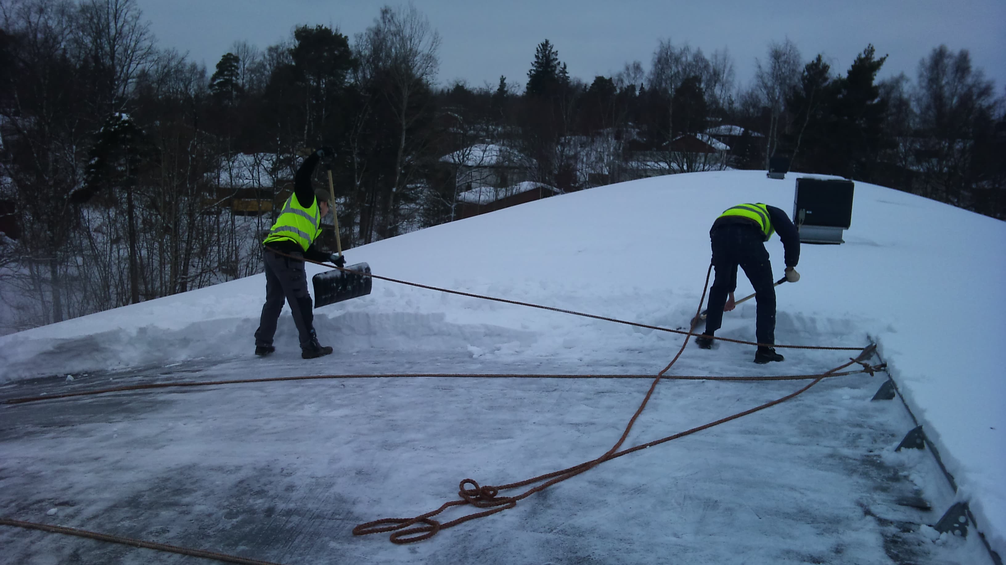 Två personer skottar snö på ett tak med rep och snöskyfflar.