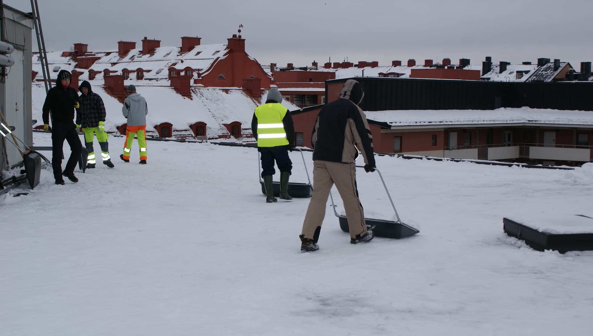 Arbetslag med snöredskap på ett snötäckt tak framför stadens takåsar.